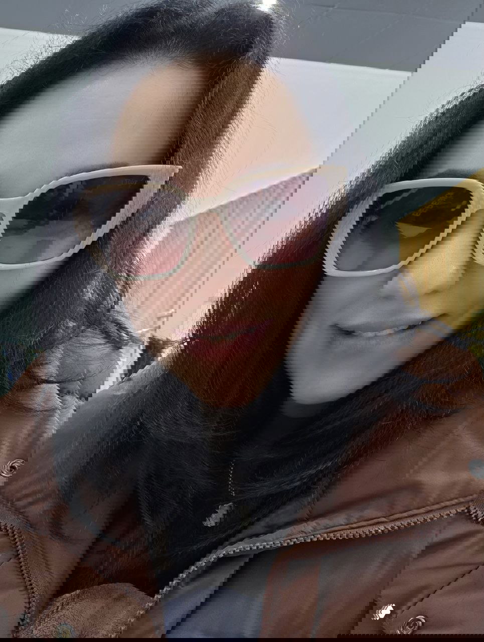 Женщина с тёмными волосами в бежевых очках и коричневой куртке улыбается в примерочной, видны элементы гардероба на заднем плане.