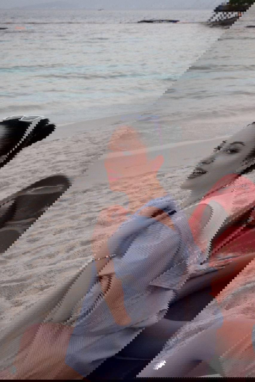 Женщина в лёгкой одежде сидит на пляже рядом с каяком, улыбается и смотрит в сторону камеры; пляжный пейзаж и расслабленная поза на закате/вечере.