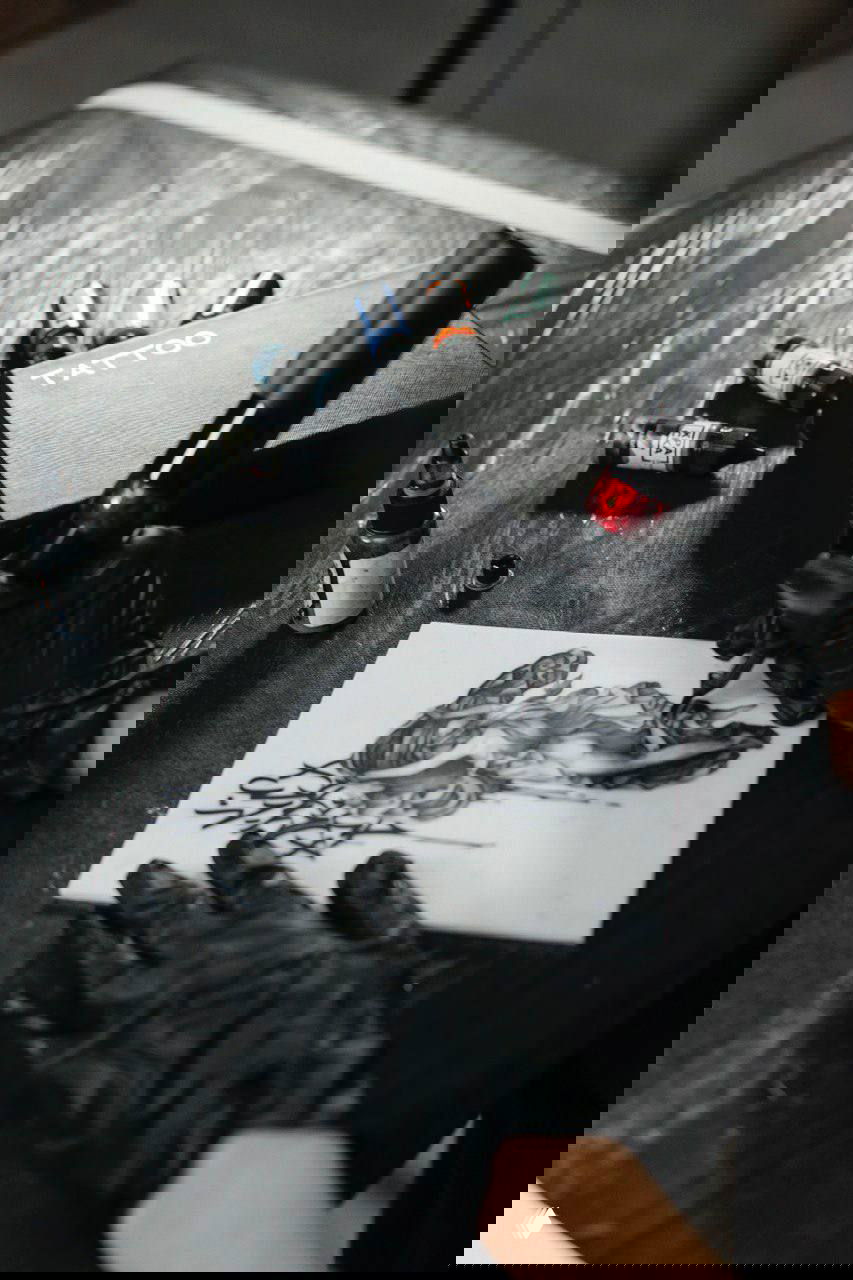 Крупный план рабочего места мастера: тату-машинка в руке, бутылочки с красками и шаблон с рисунком для практики татуировки.