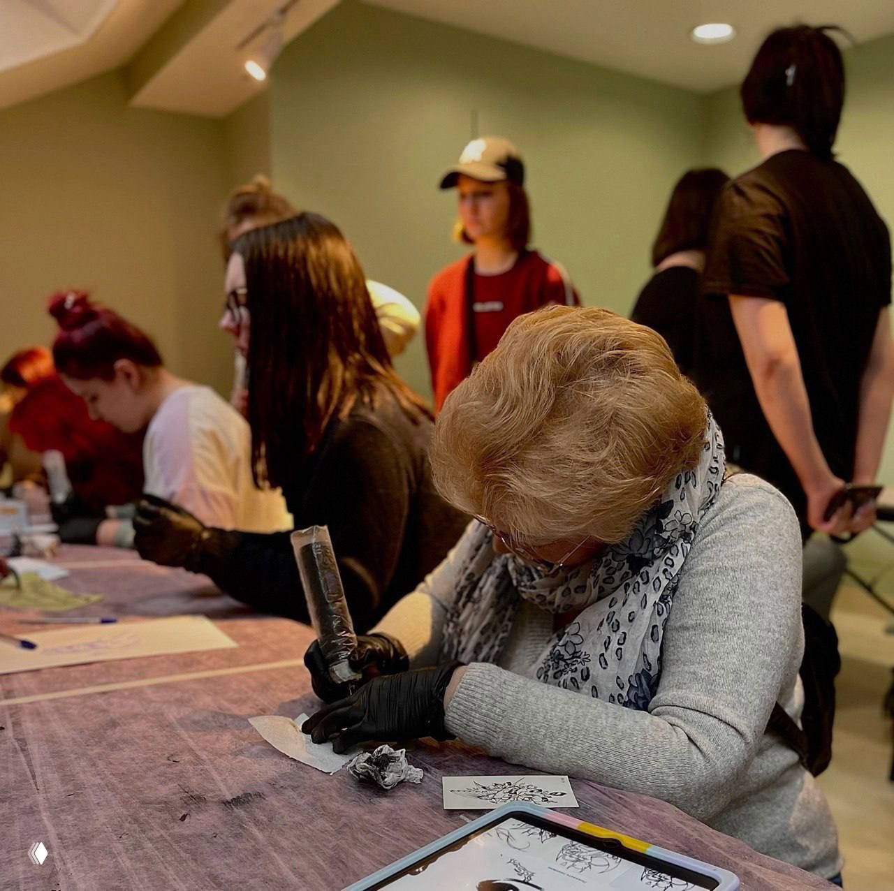Группа участников за столами на мастер-классе по тату: практические упражнения, внимательное руководство наставников и рабочие столы с материалами.