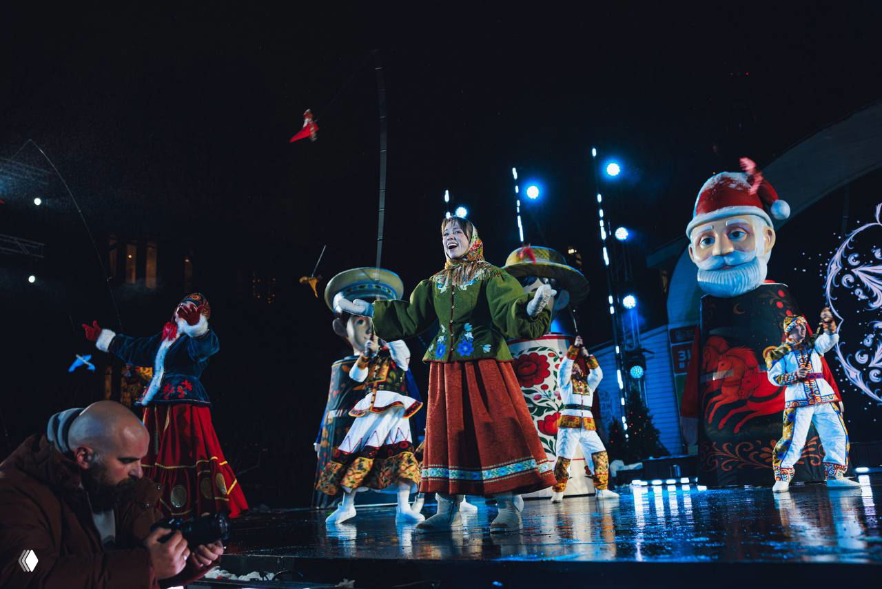 Фотограф в кадре снимает артистов на переднем плане, на заднем плане — большие расписные фигуры и артисты в традиционных нарядах.