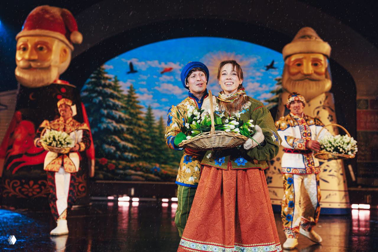 Актёр и актриса с корзинами подснежников на фоне расписных гигантских фигур месяцев — театральная иллюстрация сказки «12 месяцев».