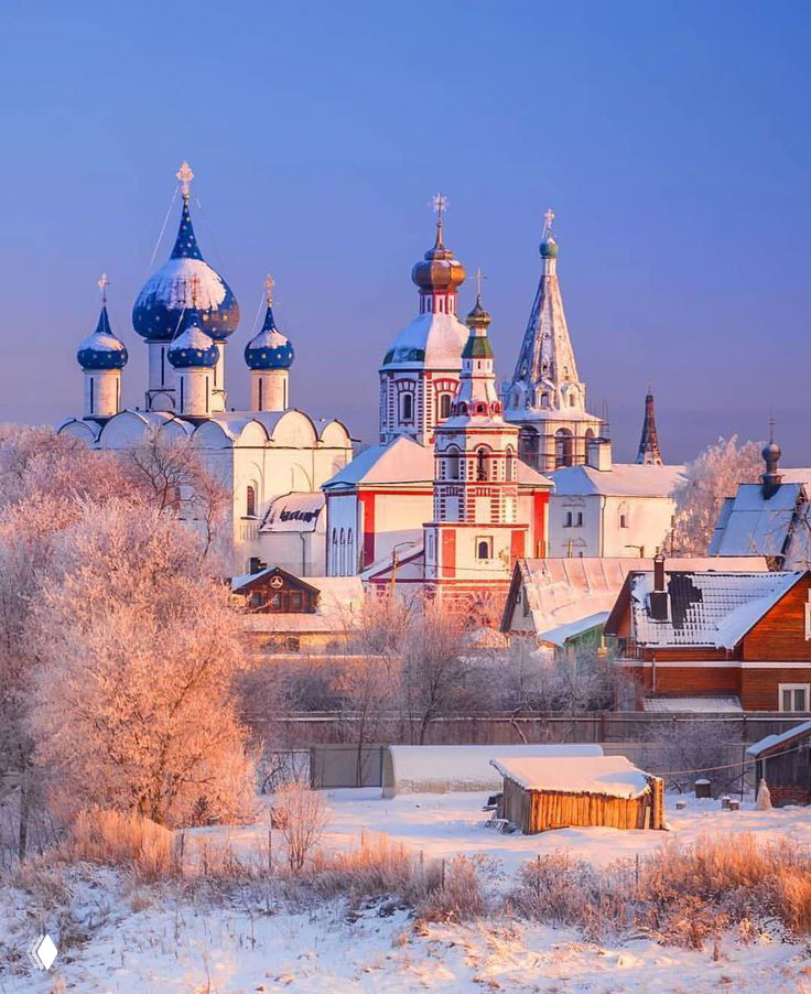 Зимний вид на Суздаль: заснеженные купола церквей и старинные постройки на фоне холодного розового света рассвета, уютная провинциальная сцена.