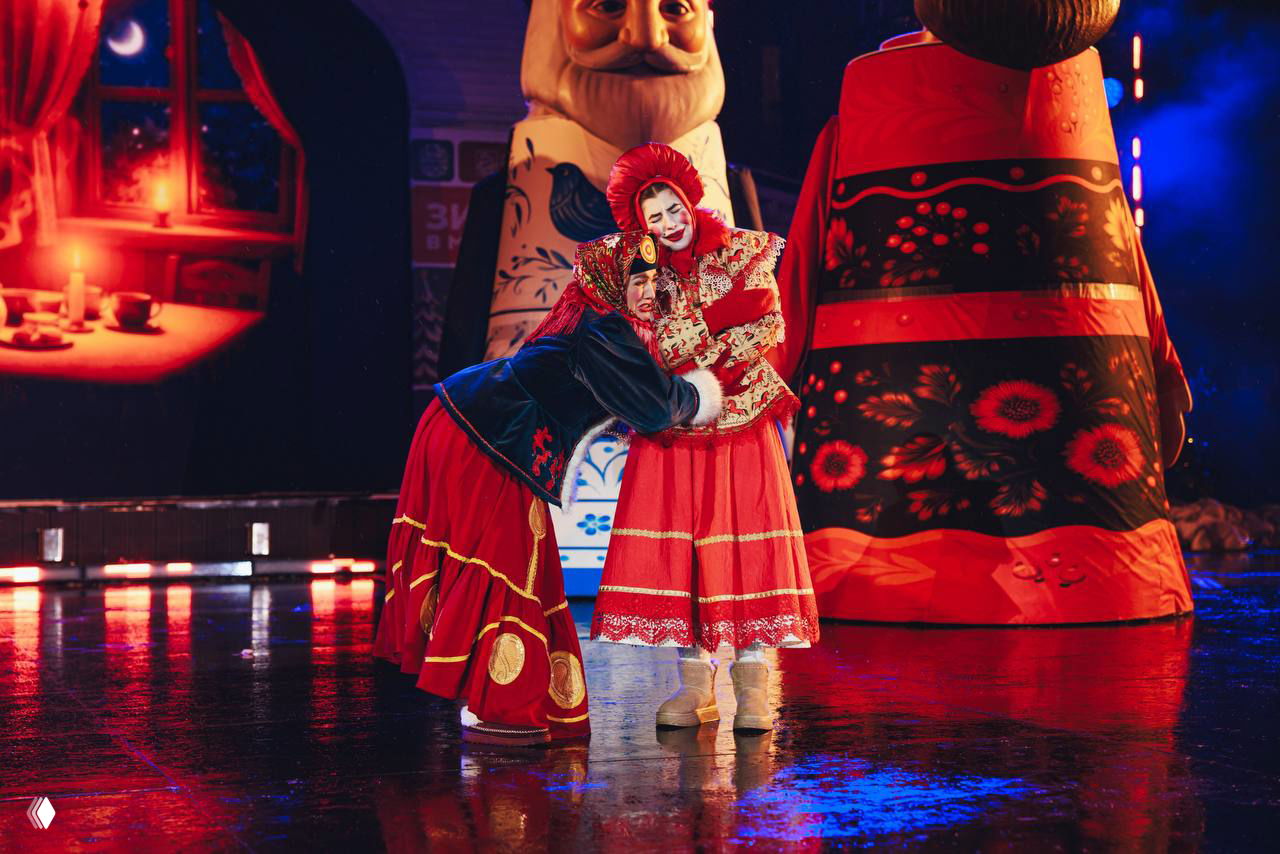 Два актёра в выразительных традиционных костюмах в центре сцены на фоне крупной декоративной фигуры и яркой подсветки.