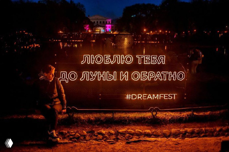 Неоновая инсталляция «Люблю тебя до Луны и обратно» у воды, тёплая подсветка и посетитель на скамейке рядом с инсталляцией.