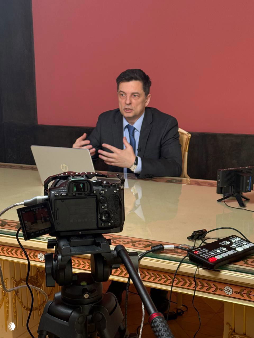 Камера и трансляционное оборудование на штативе в первом плане, на заднем плане ведущий в костюме за ноутбуком готовится к эфиру на фоне розовой стены.