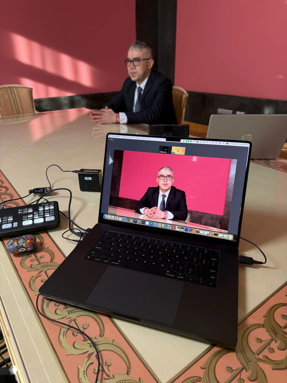 Рабочий стол с ноутбуком, на экране которого отображён удалённый спикер в костюме; рядом техническое оборудование и в фоне участник трансляции.