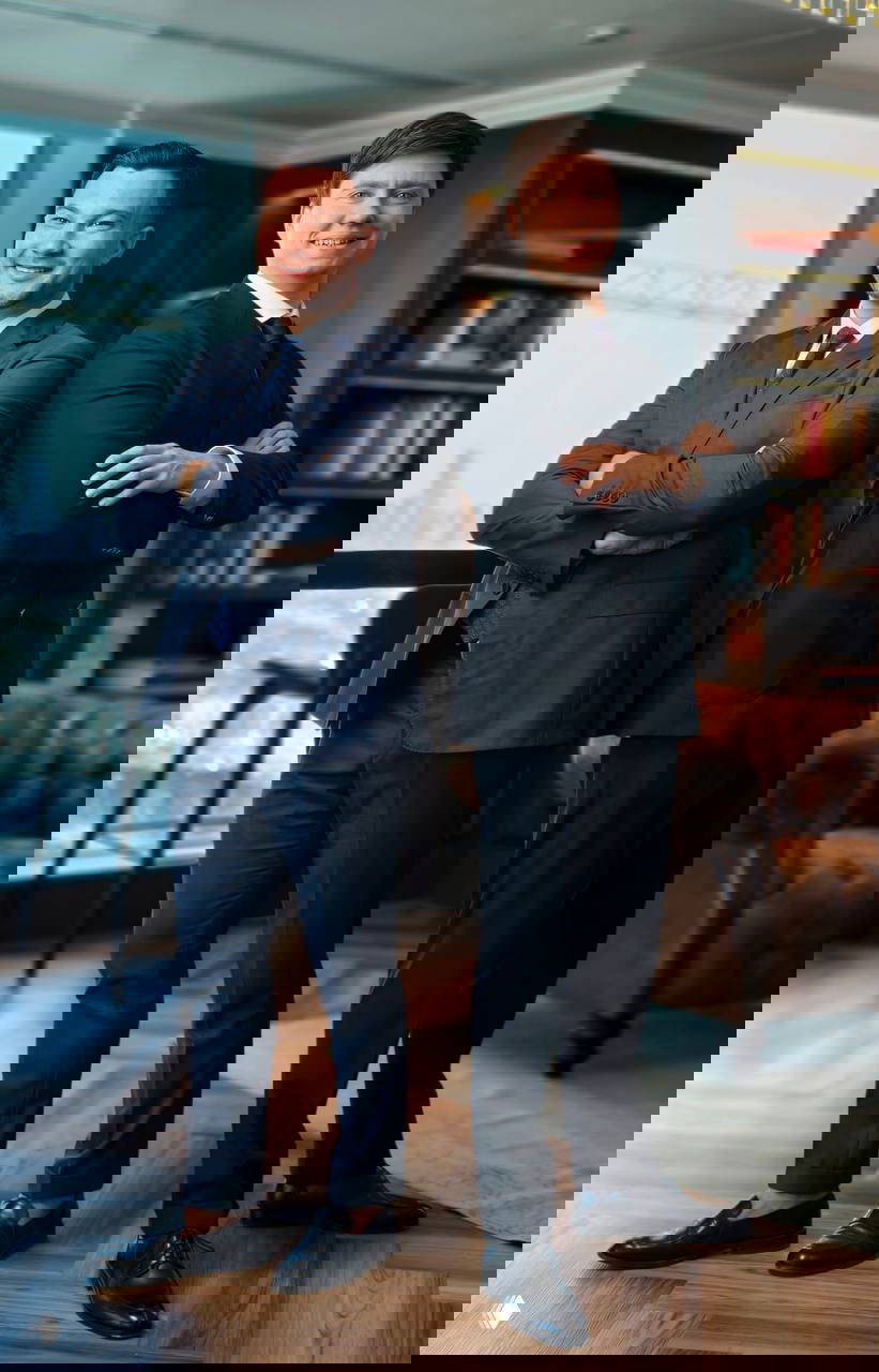 Два мужчины в деловых костюмах в офисном интерьере, стоят в полный рост на фоне окна с видом на город и книжные полки.