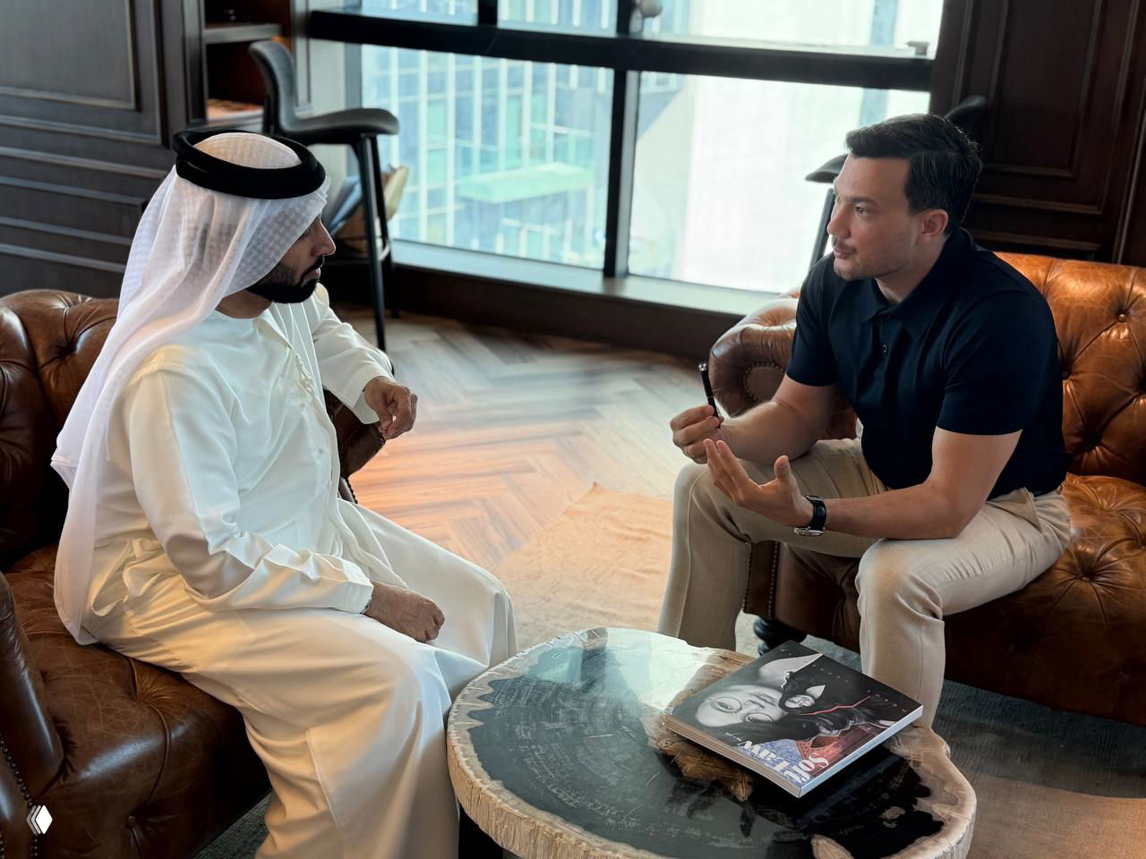 Мужчина в традиционной арабской одежде и собеседник в тёмной рубашке беседуют у журнального столика в офисе с видом на город.