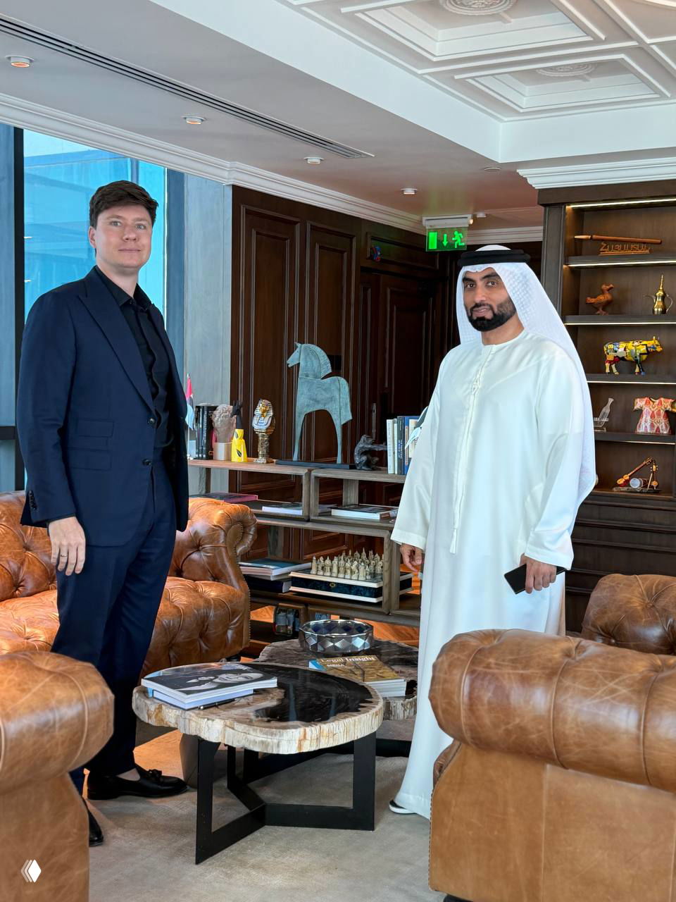 Двое мужчин — один в деловом костюме, другой в традиционной арабской одежде — стоят в элегантном деревянно-кожаном офисе рядом с дизайнерской мебелью.