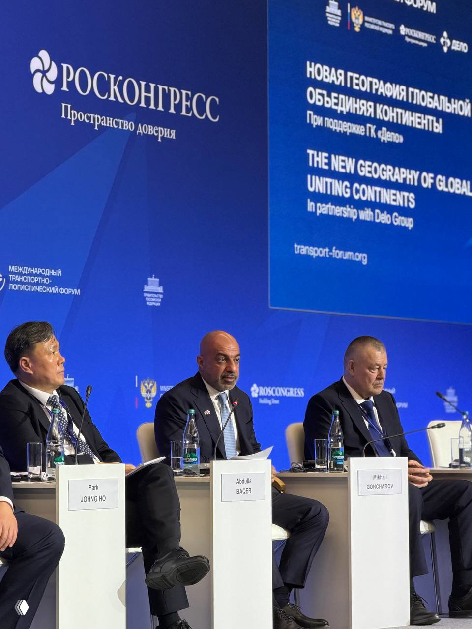 Панельная дискуссия на сцене форума: несколько делегатов сидят за столом, на столах таблички с именами и бутылки воды, синий фон с брендингом мероприятия.