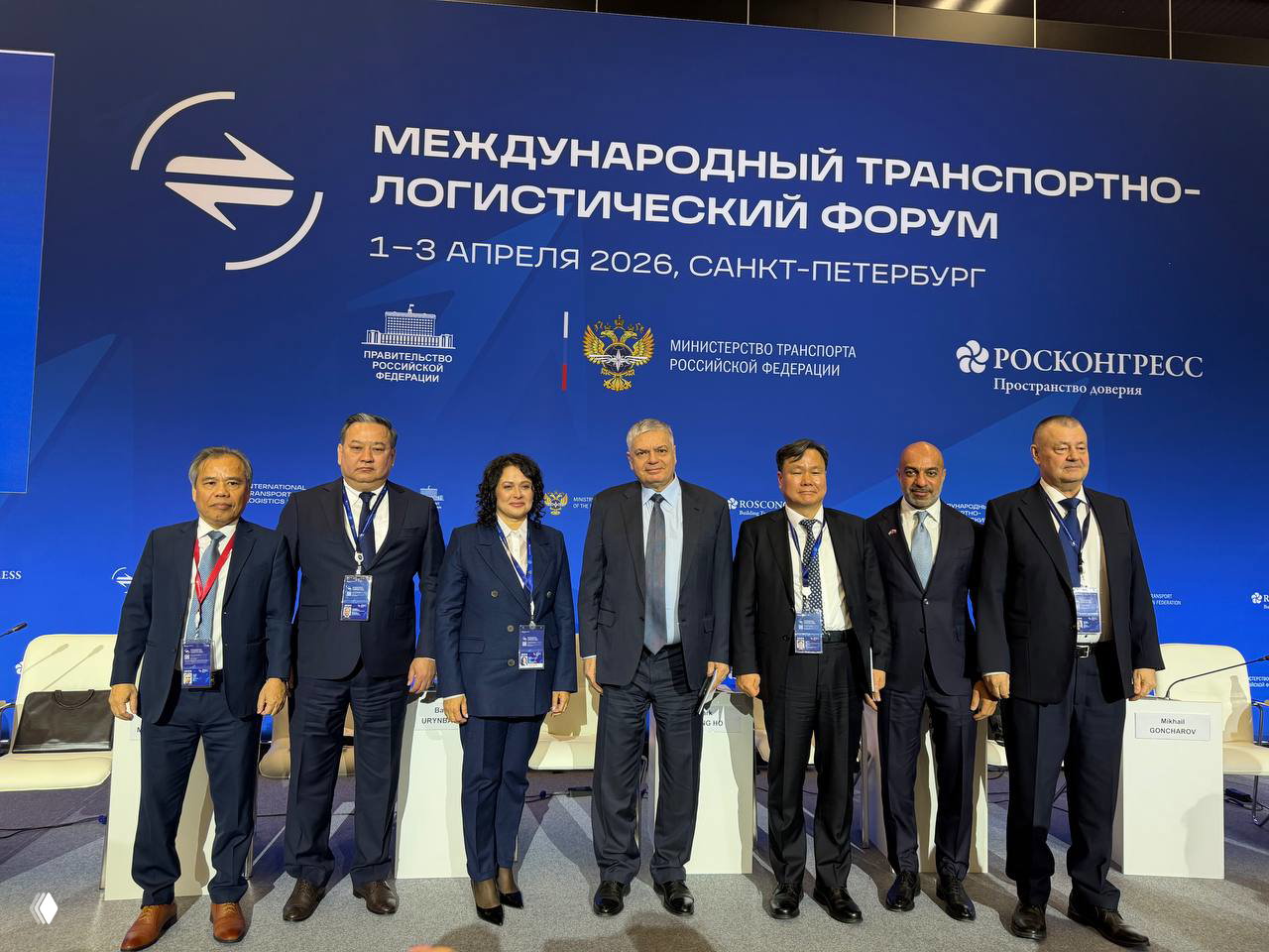 Групповая фотография делегатов на фоне большого синего баннера форума: официальные представители стоят в деловой одежде у брендинга мероприятия.