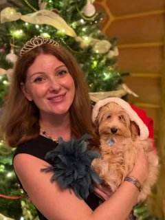 Женщина улыбается, держит маленького пушистого щенка на руках на фоне украшенной новогодней ёлки — тёплая семейная сцена, праздничный отдых.