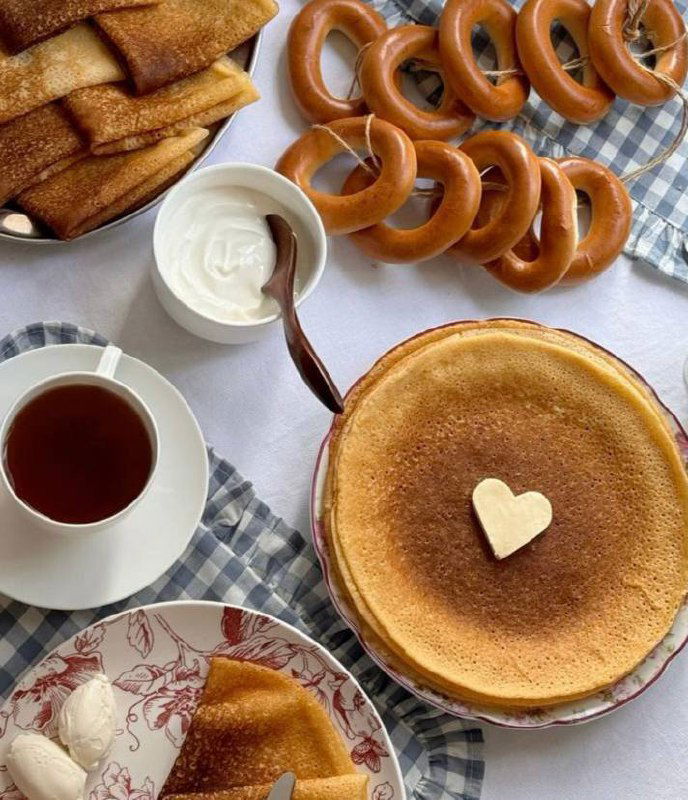 Тарелки с блинами и чашки на столе; изображение частично искажено цифровыми артефактами, видны блинчики и сервировка.