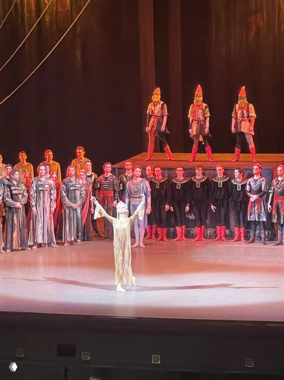 Крупный план на сцене: прима-балерина в центре с поднятыми руками, вокруг — ансамбль артистов в костюмах спектакля.