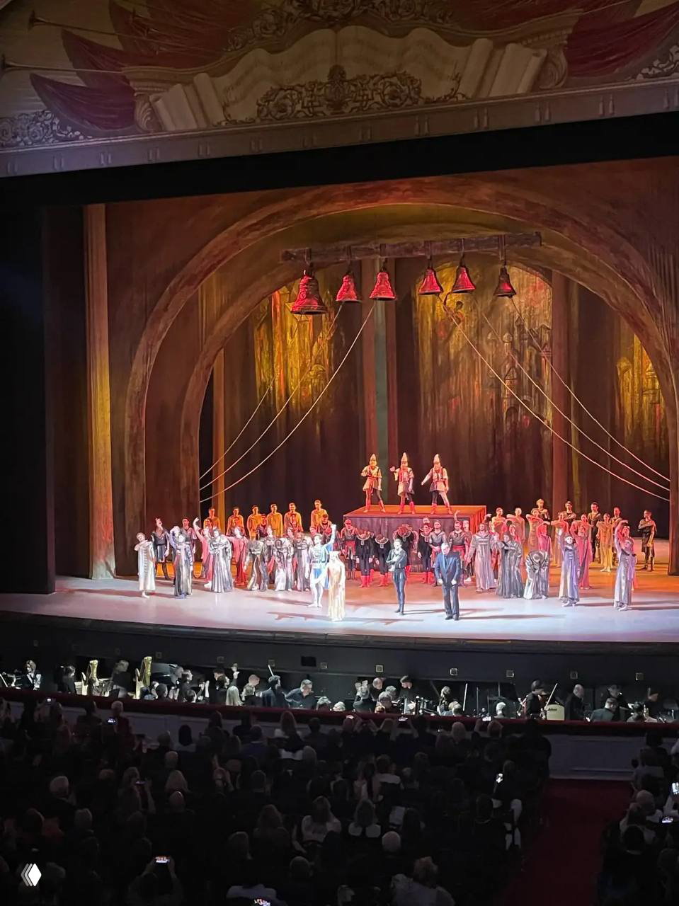 Широкий кадр постановки на сцене с арочной декорацией и подвешенными колоколами, на сцене много исполнителей.