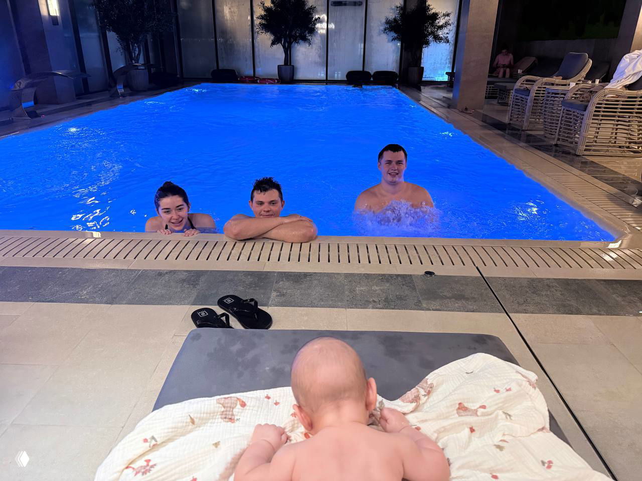 Внутренний бассейн: трое взрослых находятся в воде у бортика, на переднем плане видна голова и спина младенца на лежаке — семейный отдых в отеле.