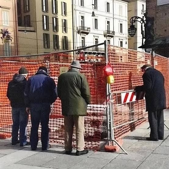 Группа пожилых мужчин стоит у оранжевого строительного забора в Италии, сосредоточенно наблюдая за проводимыми работами и разговаривая между собой.