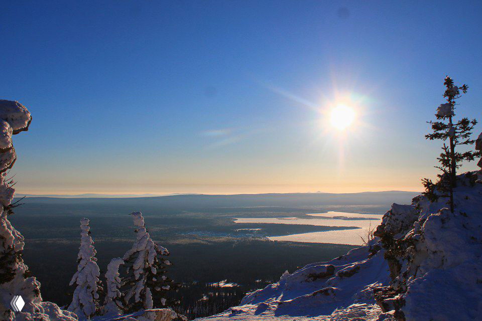 Широкий панорамный вид с вершины: солнце, долины, извилистые берега озера и снежные склоны — пейзаж походного маршрута в Зюраткуле.