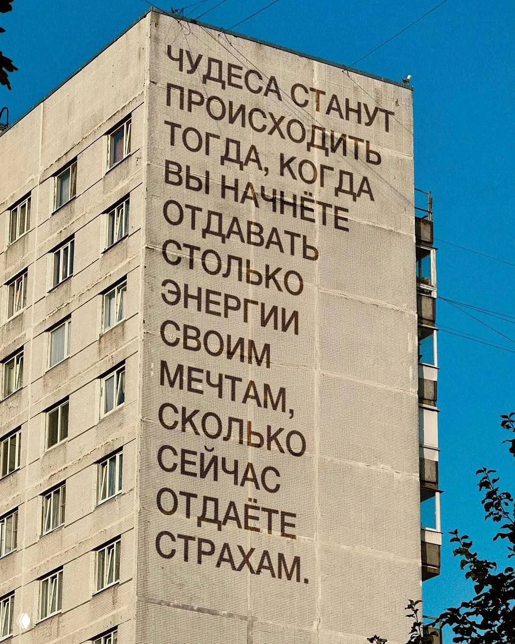 Мотивирующая надпись на стене жилого дома: текст о чудесах и страхах, фотография крупным планом на фоне ясного неба и городской среды.