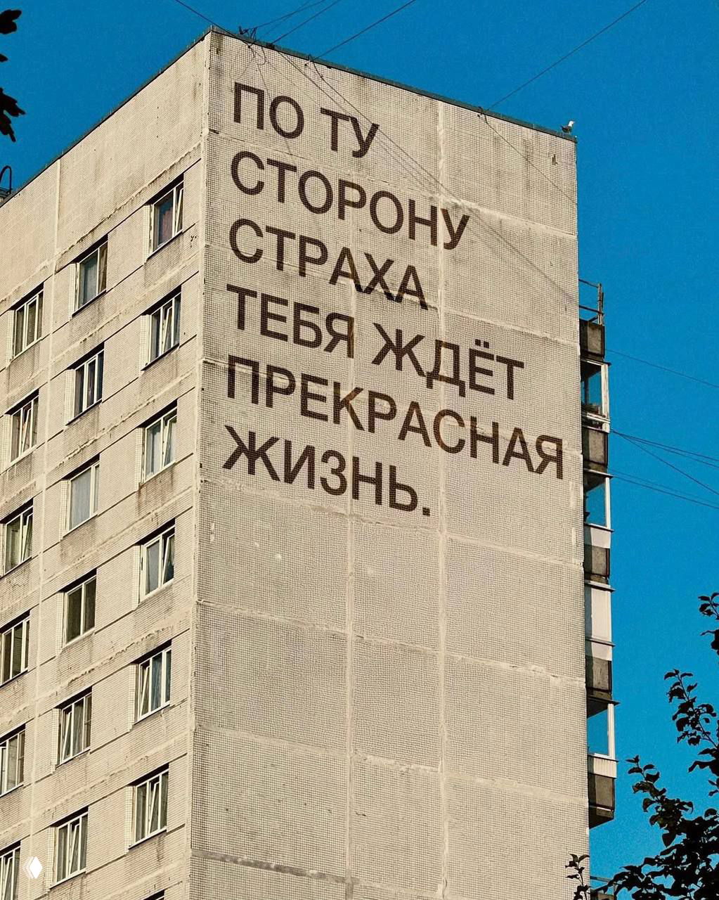 Фасад высотного дома с надписью: «По ту сторону страха тебя ждет прекрасная жизнь», мотивирующий текст на фоне синего неба и веток деревьев.