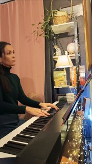 Женщина играет на электронном фортепиано в домашнем кабинете; видна лампа, полки с предметами и гирлянда — иллюстрация музыцирования и контекста обсуждения памяти.