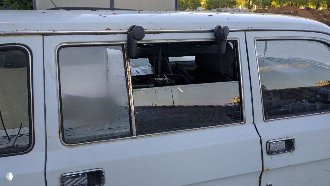 Боковая часть старого белого автомобиля с отсутствующим стеклом в боковой двери, заметны ржавчина, отражения и фрагменты салона внутри.