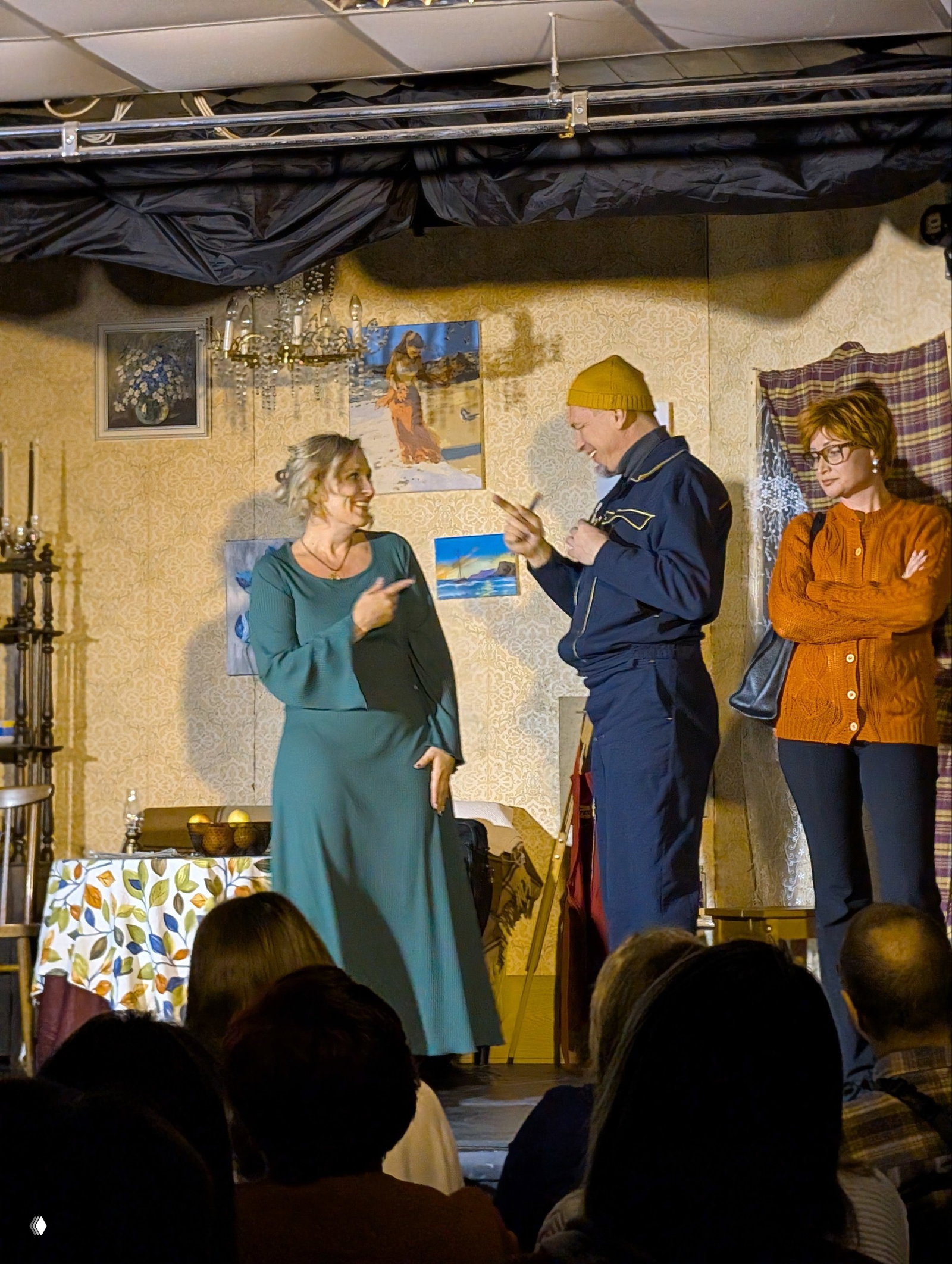 Сцена с тремя актёрами: женщина в зелёном платье улыбается, мужчина в рабочей форме жестами отвечает, другая актриса наблюдает с рук на груди.