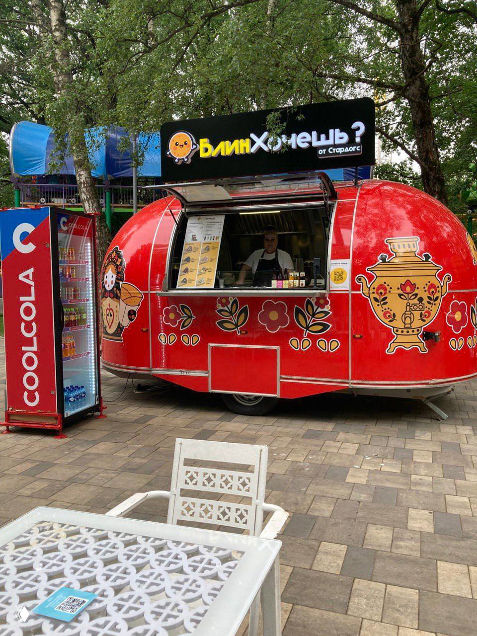 Красный фудтрак «БлинХочешь?» от Stardogs в парке развлечений в Химках, украшен народными узорами и изображением самовара, видно окошко продаж и столик в первом плане.