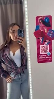 Женщина делает зеркало‑селфи в бытовке; на зеркале видна яркая рекламная наклейка Ozon с логотипом и промо‑графикой, отражение интерьера частично видно.