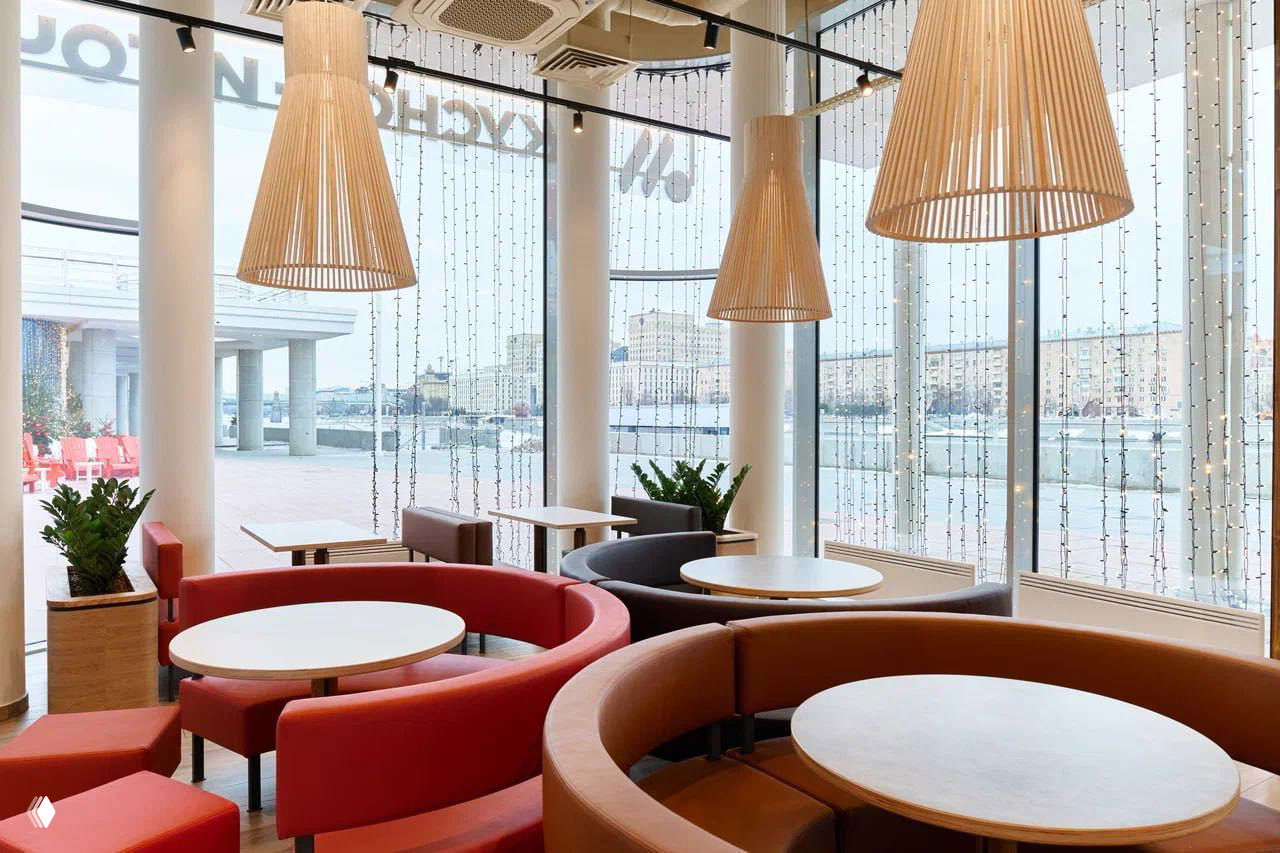 Зона посадки с круглыми диванами и столами, дизайнерские подвесные светильники и большие окна ресторана «Вкусно — и точка», вид на реку и будущую веранду.