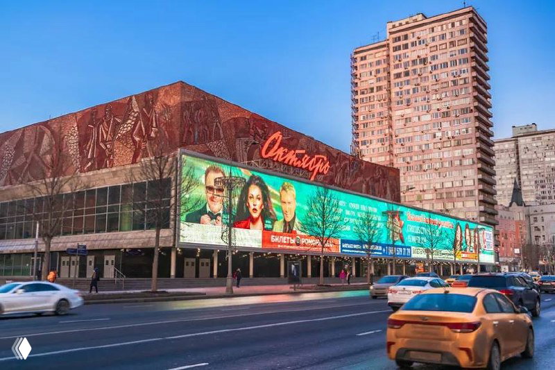 Фотография существующего фасада кинотеатра «Октябрь» на Новом Арбате с неоновой вывеской, рекламной лентой и уличным движением на переднем плане днем.