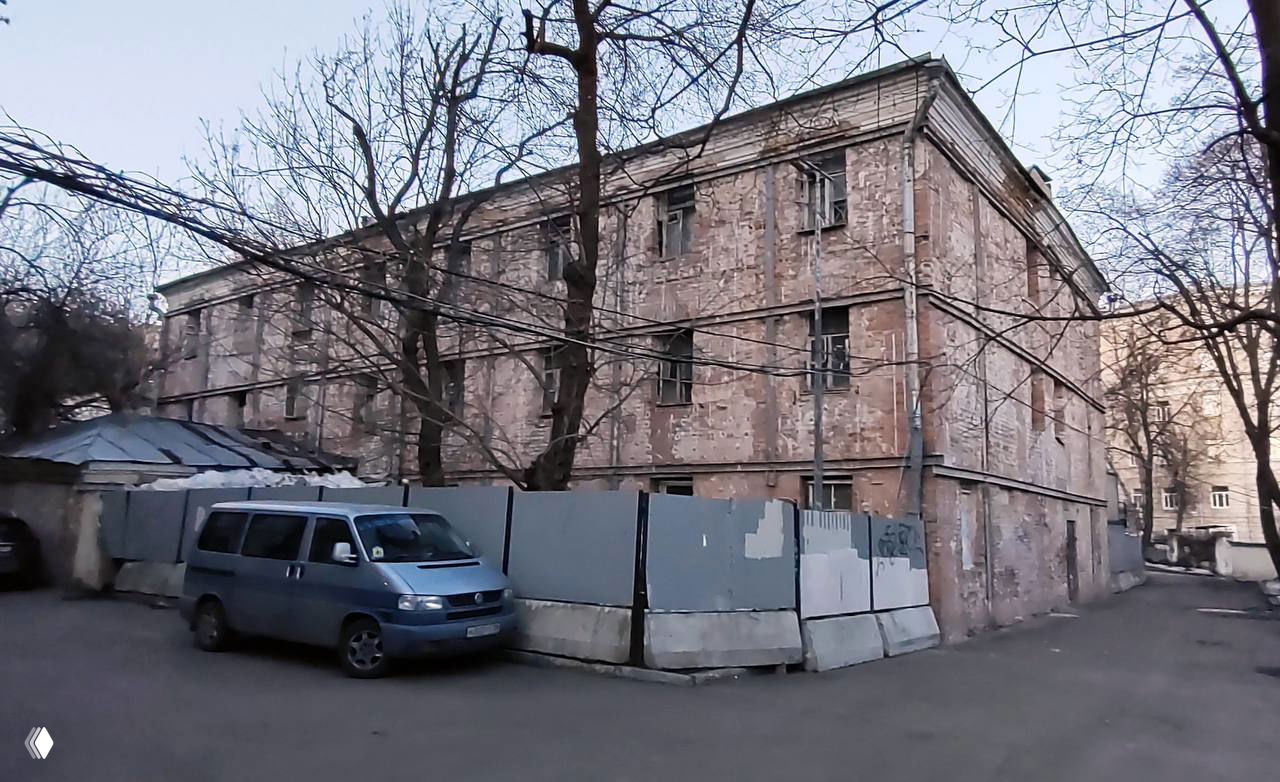 Запущенное здание усадьбы до реставрации: облупившаяся кирпичная кладка, временный забор, нависающие провода и припаркованный фургон.