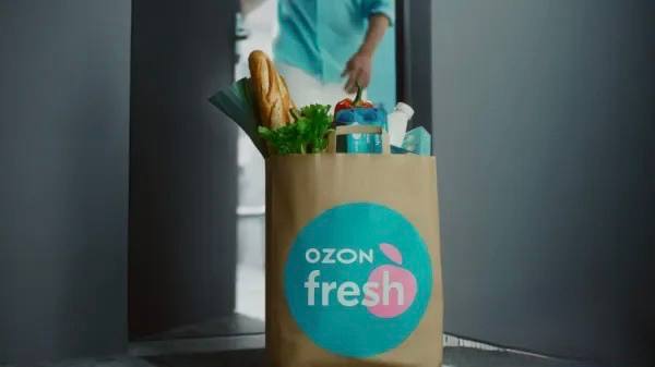Бумажный пакет с логотипом Ozon fresh и продуктами (хлеб, зелень, упаковки) на пороге — иллюстрация сервиса 30‑минутной доставки и концепции дарксторов в городе.