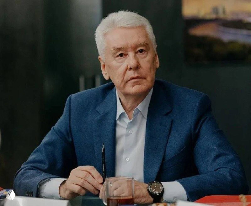 Портрет пожилого мужчины в синем пиджаке за столом с ручкой в руках и стаканом, деловая обстановка, без указания личности.