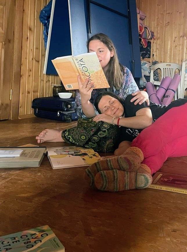 Уютная сцена в деревянном зале: участницы лежат и читают вслух, вокруг книги и мягкие опоры, ощущение близости и практики в группе.
