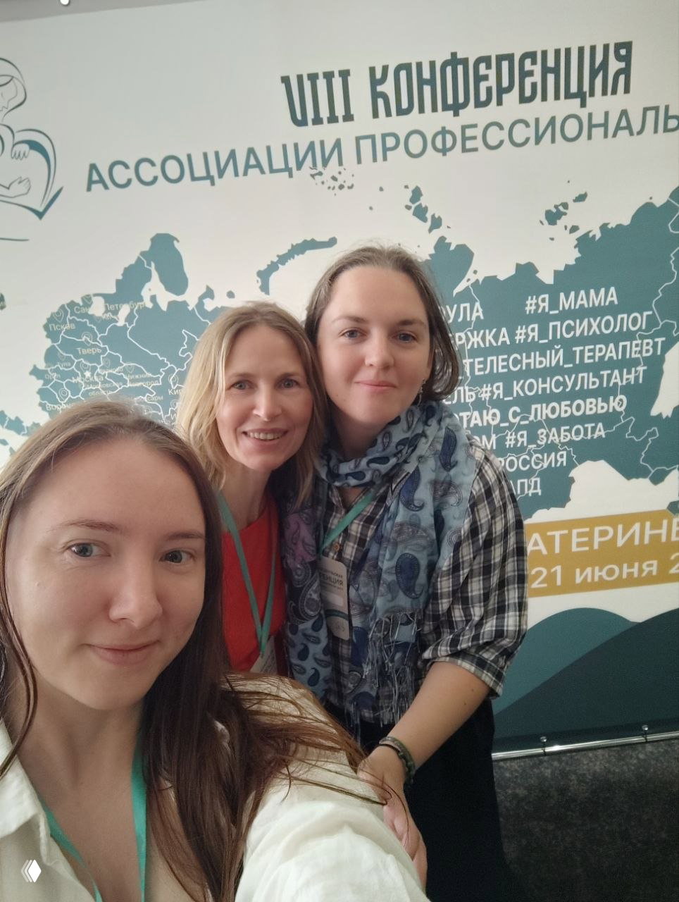 Селфи трёх участниц на фоне баннера VIII конференции Ассоциации Профессиональных Доул, видно карту и текст мероприятия, у всех бейджи и тёплые улыбки в конференц-пространстве.