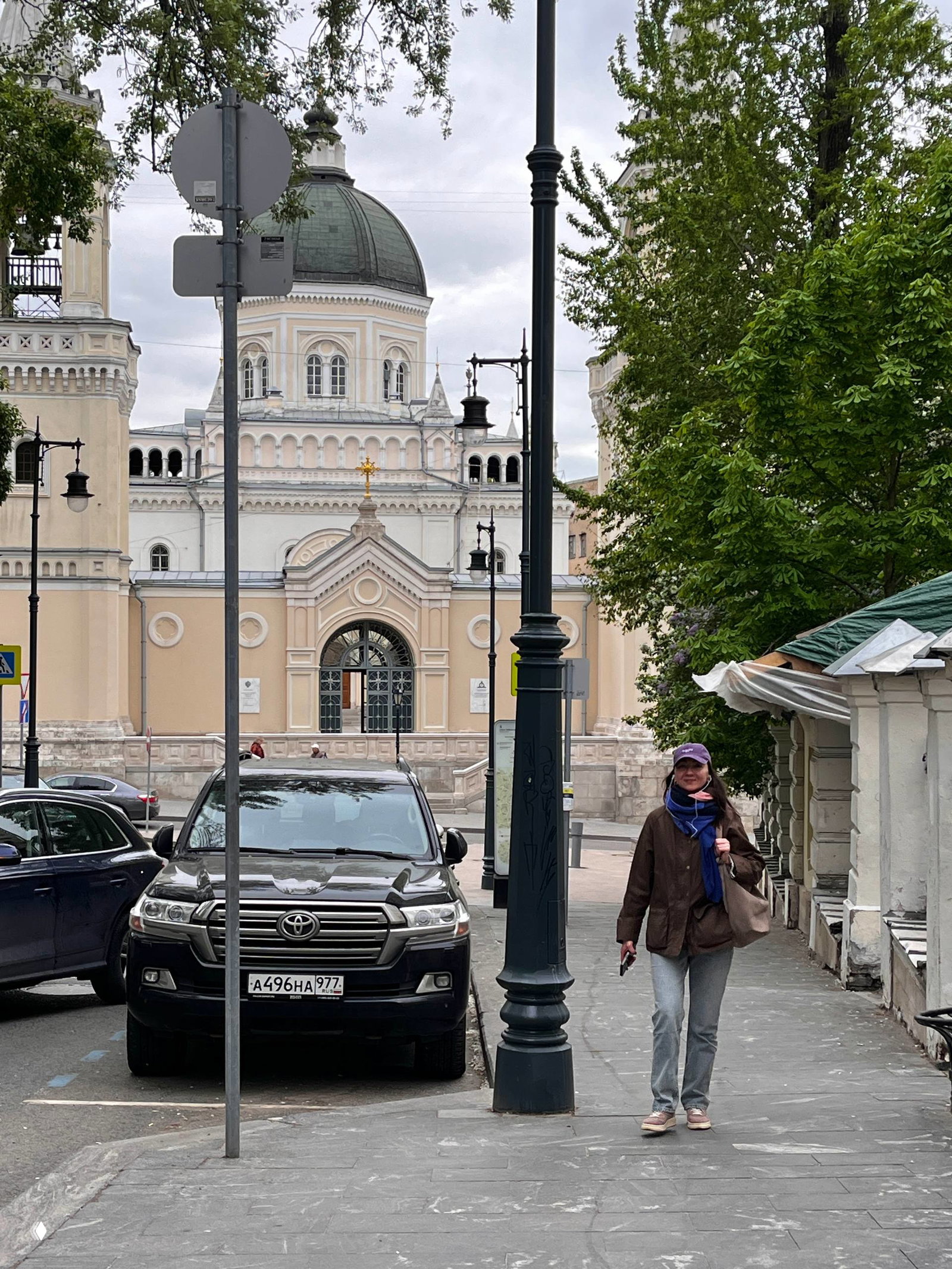 Улица с куполом и фасадом храма на дальнем плане, припаркованный автомобиль и прохожая — городской пейзаж маршрута по центру Москвы.