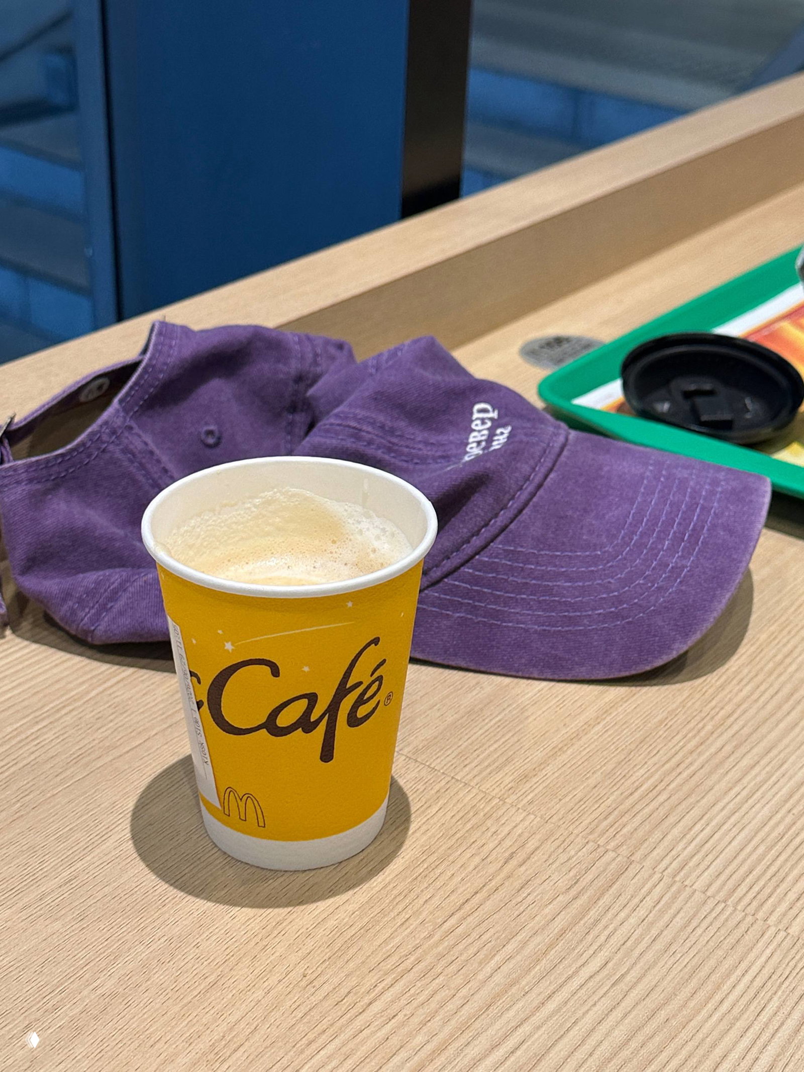 Крупный план: бумажный стакан McCafé с маленьким капучино на деревянном столе McDonald's; рядом фиолетовая кепка и поднос с крышкой.
