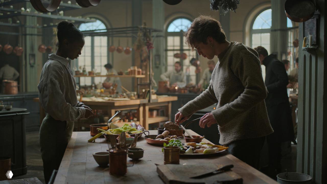 Кадр с кухонным процессом: повар и помощница работают за длинным столом, медные ковши и традиционный инвентарь создают атмосферу эпохи.