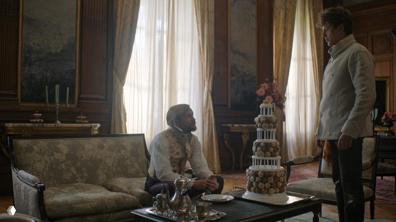 Диалоговая сцена в салоне: двое мужчин и высокий торт из пирожных на столе, роскошная мебель и свет из окон подчеркивают декор.