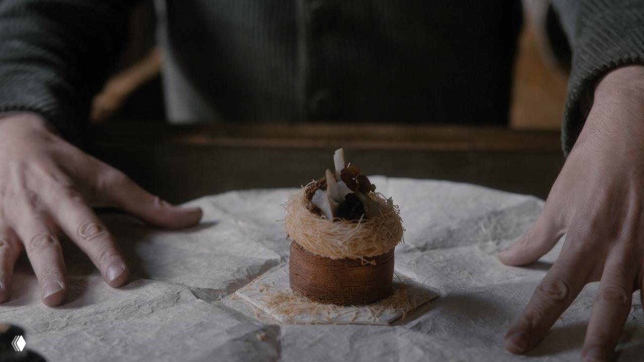 Миниатюрный сложный десерт на столе, оформленный как мини-скульптура: корж, нитевидная посыпка и декоративные элементы, эстетика подачи.