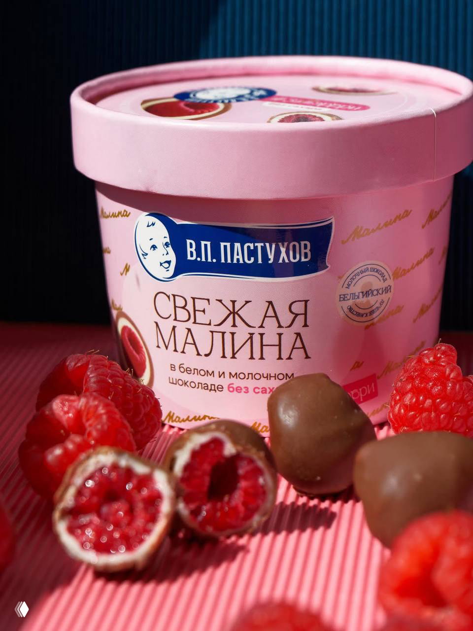 Крупный план банки с маркировкой В.П. Пастухов «Свежая малина», перед ней разрезанные ягоды и шоколадные шарики на розовой подложке — пример освещения и композиции.