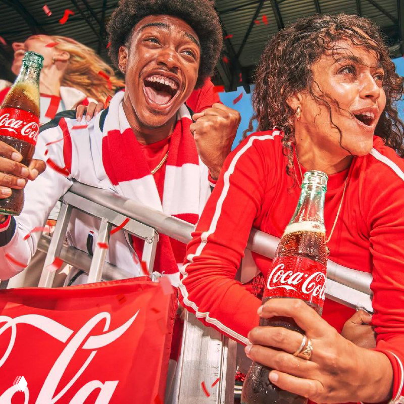 Восторженные фанаты кричат и держат бутылки Coca‑Cola у ограждения трибуны — кадр с ярким красным оформлением и конфетти.