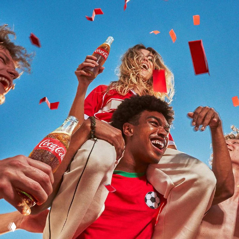 Парень с девушкой на плечах держит бутылку Coca‑Cola на фоне голубого неба — праздничный кадр с конфетти и чувством триумфа на стадионе.