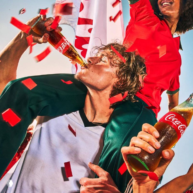 Болельщик пьет Coca‑Cola среди летящих красных конфетти — динамичная сцена рекламной съемки в стилистике спортивного праздника.