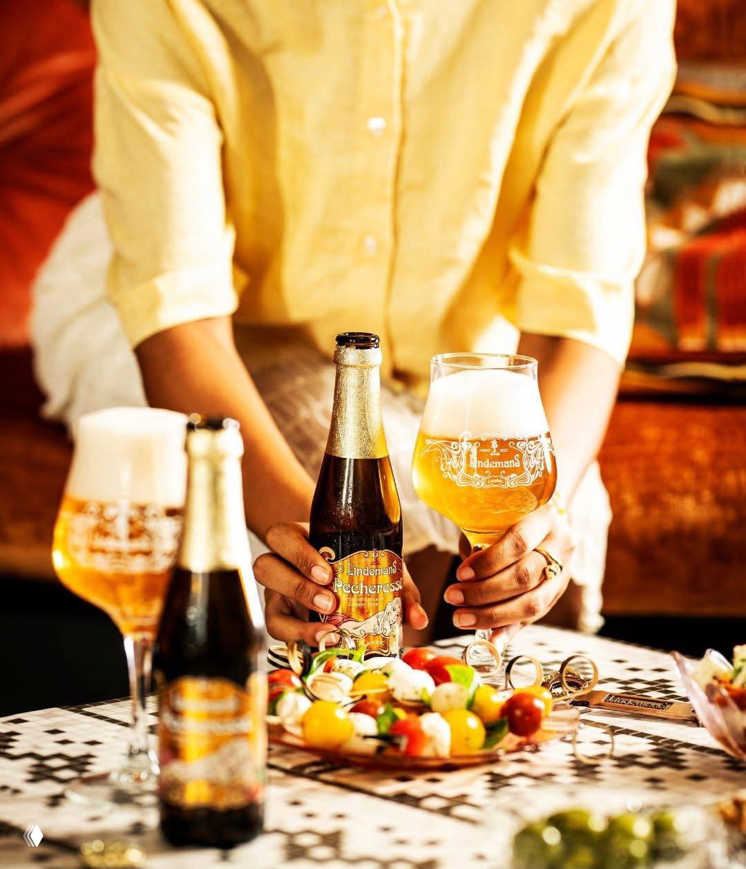 Рука держит бутылку Lindemans и наливает в бокал на фоне закусок на шпажках; тёплые тона, мягкий свет и фокус на бутылке и бокале.