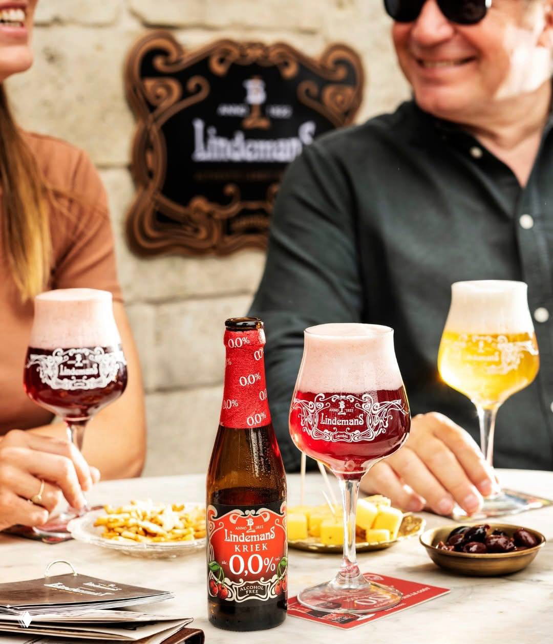 Красные малиновые ламбики и бутылка Lindemans 0,0% на столе с сыром и оливками, люди на заднем плане улыбаются — честная гастрономия без перегруза.