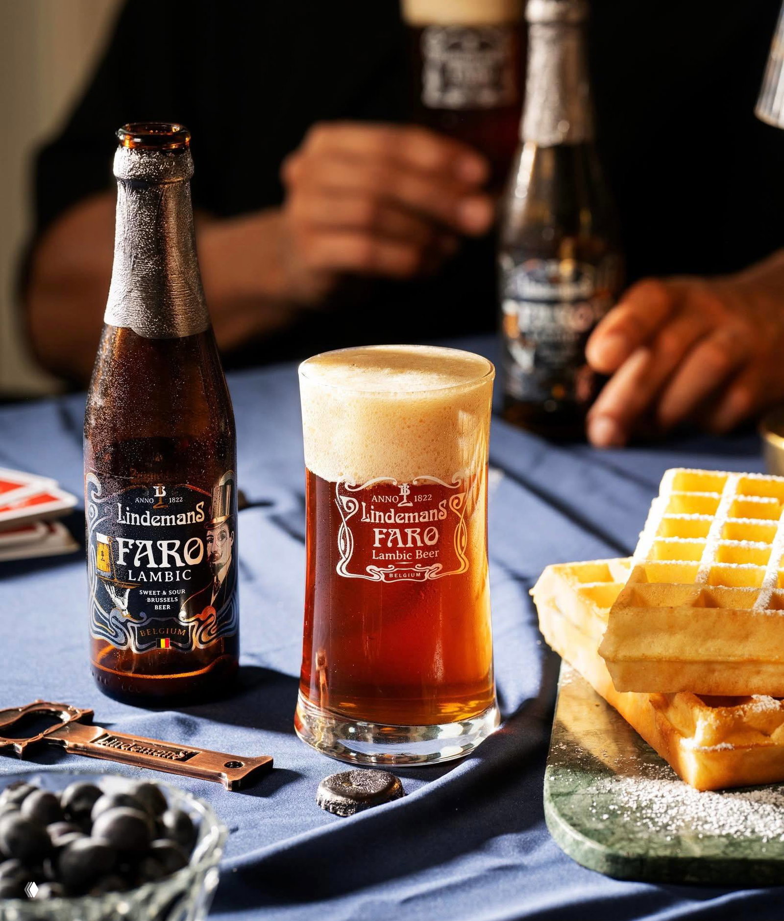 Крупный план: бутылка Lindemans FARO и высокий бокал с ламбиком рядом с вафлями на скатерти — текстура, контраст и свет подчёркивают продукт.