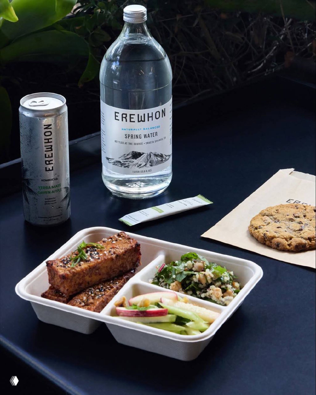 Ланч из готовых блюд Erewhon: трёхсекционный эко‑контейнер с салатом, основным блюдом и закусками, рядом бутылка воды и печенье на столе.
